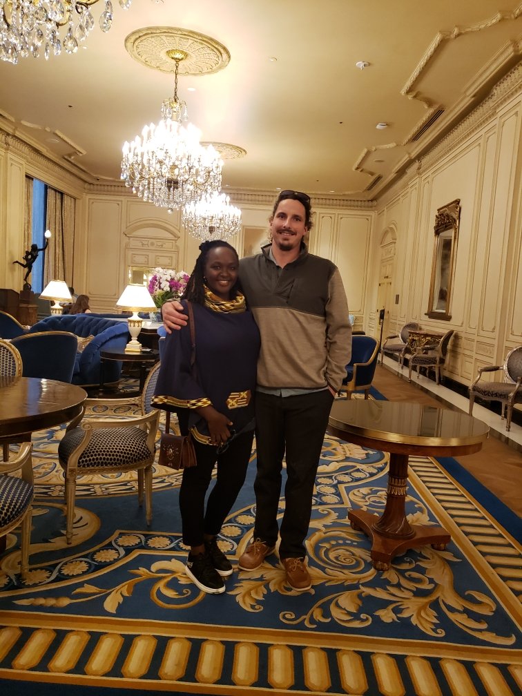 Two people smiling in an ornate room with chandeliers, elegant furniture, and patterned carpet.