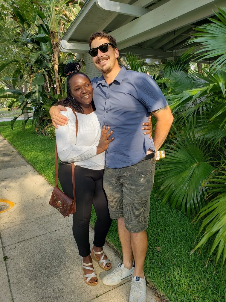 A couple posing outdoors in a garden setting with lush greenery. The woman, wearing a white top and black pants, has a brown shoulder bag and is smiling. The man, wearing sunglasses, a blue shirt, and camo shorts, stands with his arm around her. They
