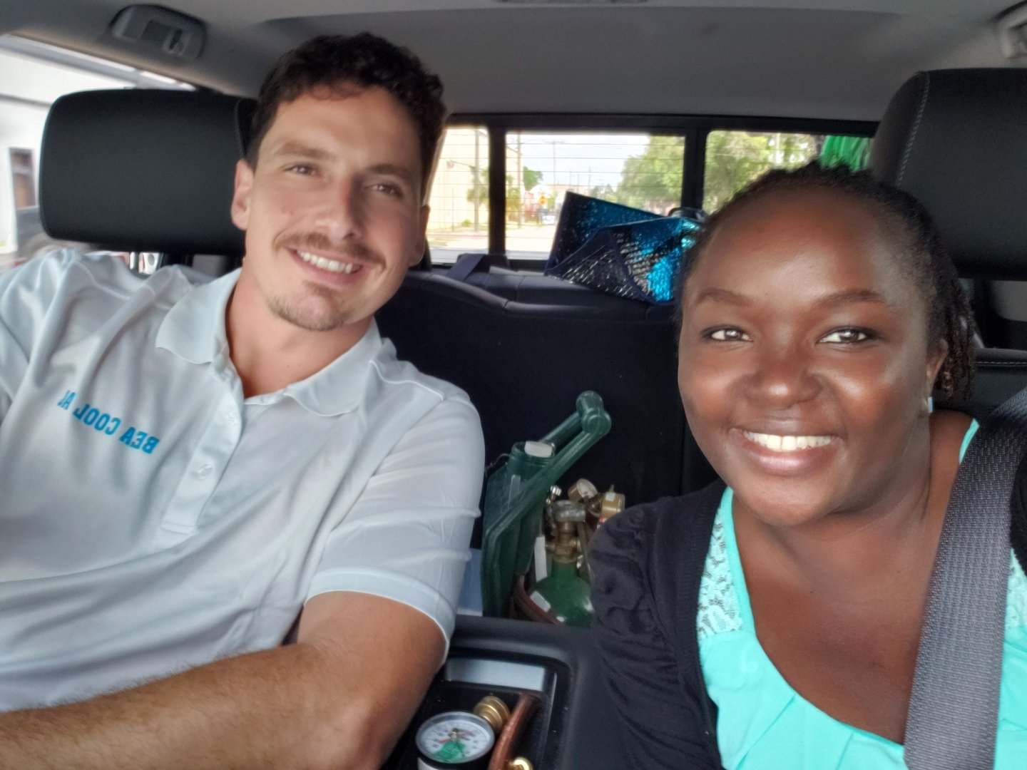 Two people smiling in a car, one wearing a white shirt and the other in a turquoise top, with various items in the backseat.