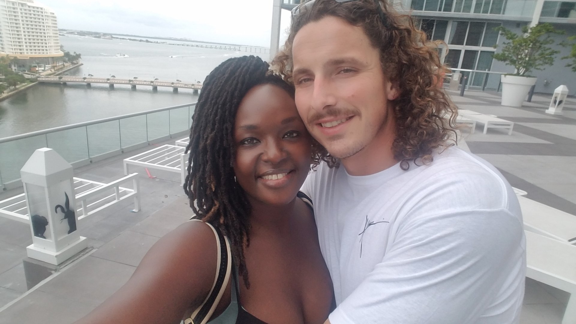 A smiling couple taking a selfie on a waterfront balcony with a view of an urban landscape and a bridge in the background.