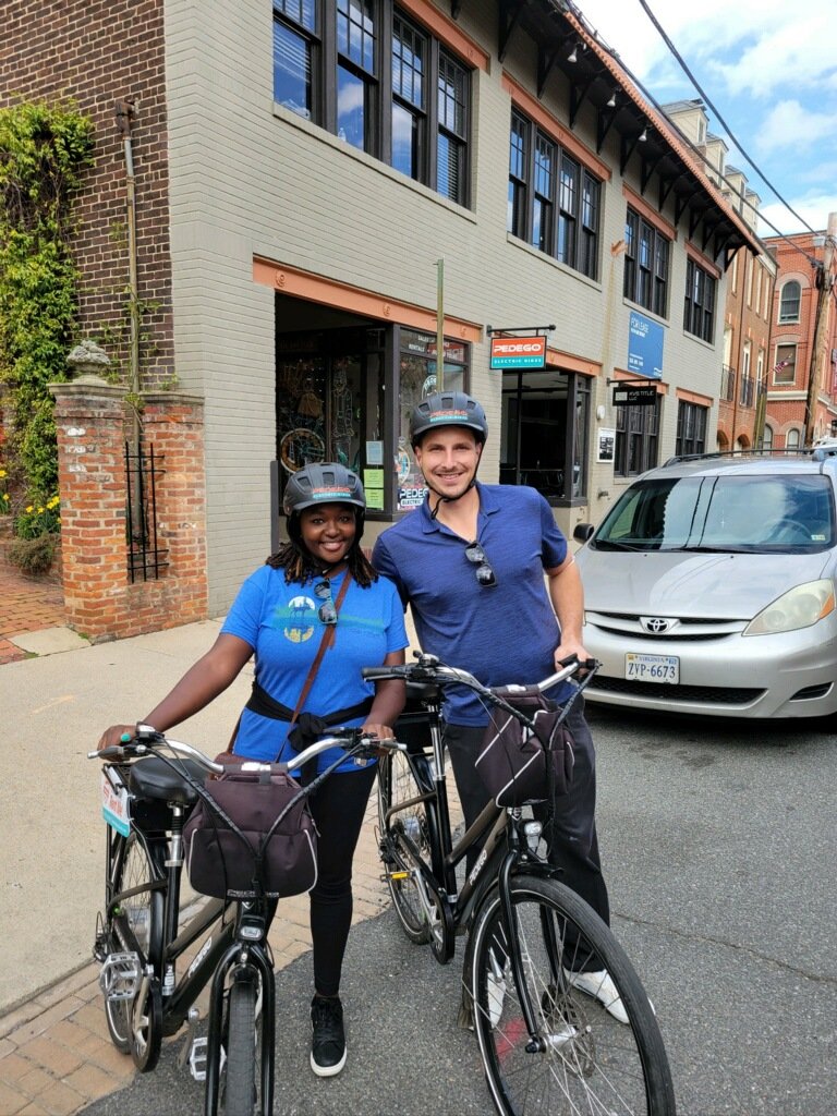 Two people wearing helmets standing with bicycles on a street in front of a brick building. A parked car and storefronts are visible in the background.