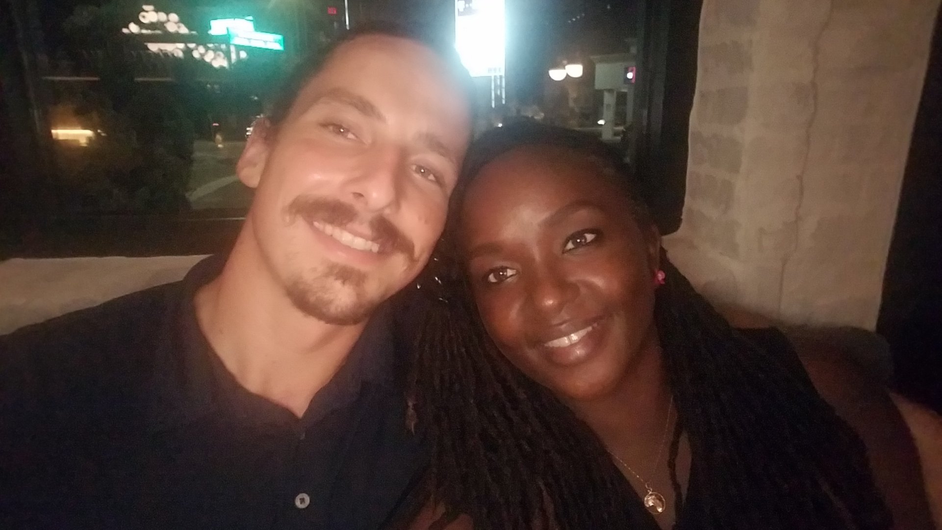 A smiling couple sitting together in a dimly lit setting, with a street scene visible through a window behind them.