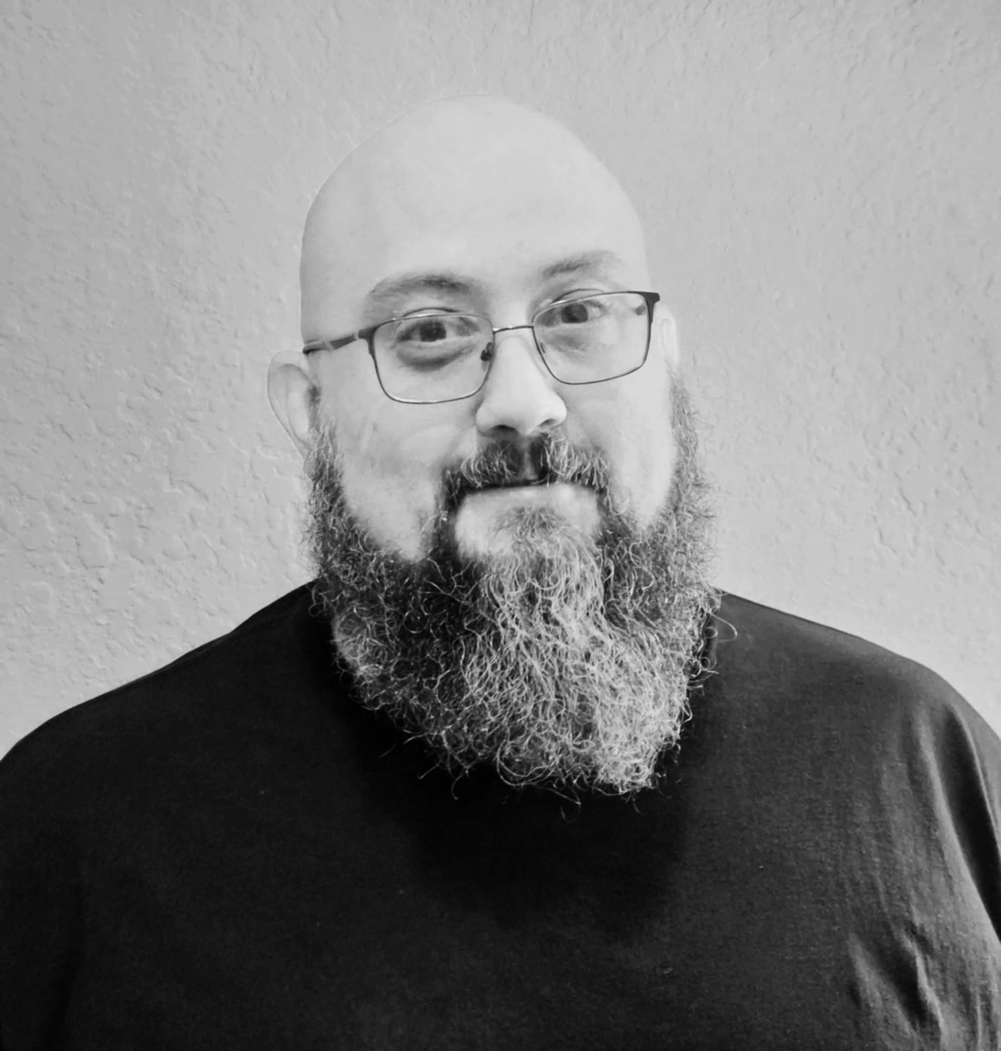 Bald man with glasses and a long beard wearing a black shirt, standing against a plain wall.