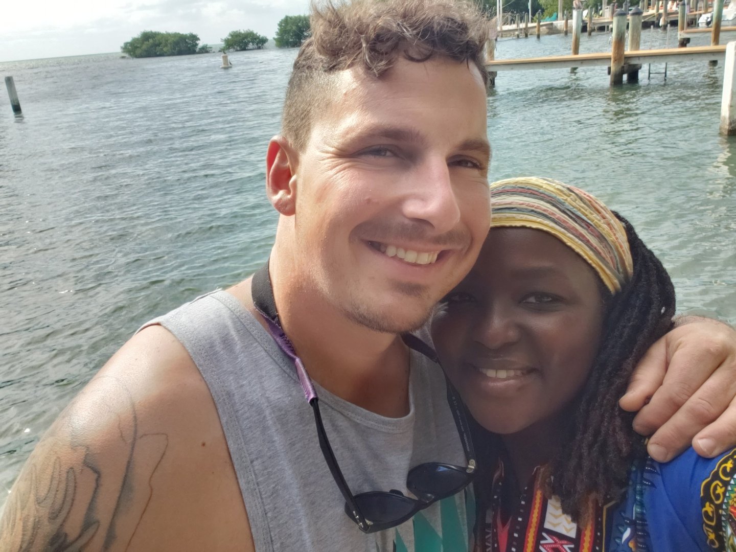 A man and a woman smiling by the waterfront with a view of the ocean and a dock in the background. The man has curly hair and is wearing a gray sleeveless shirt with sunglasses around his neck. The woman has braided hair and is wearing a colorful hea