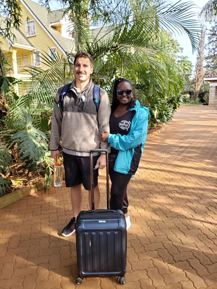 A man and a woman stand together on a brick path outside a house. The man is wearing a gray sweatshirt and black shorts, with a backpack, holding a clear water bottle, and has a black suitcase in front of him. The woman is wearing sunglasses, a blue