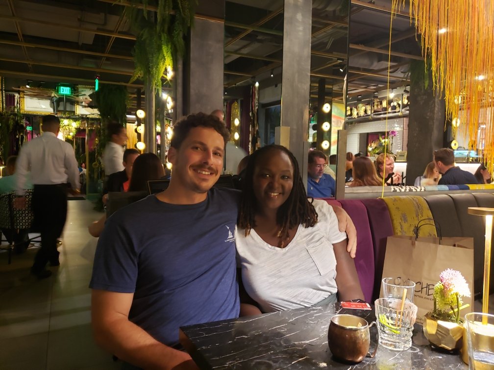 A couple sitting together at a restaurant, surrounded by other diners and decorative lighting, smiling at the camera.