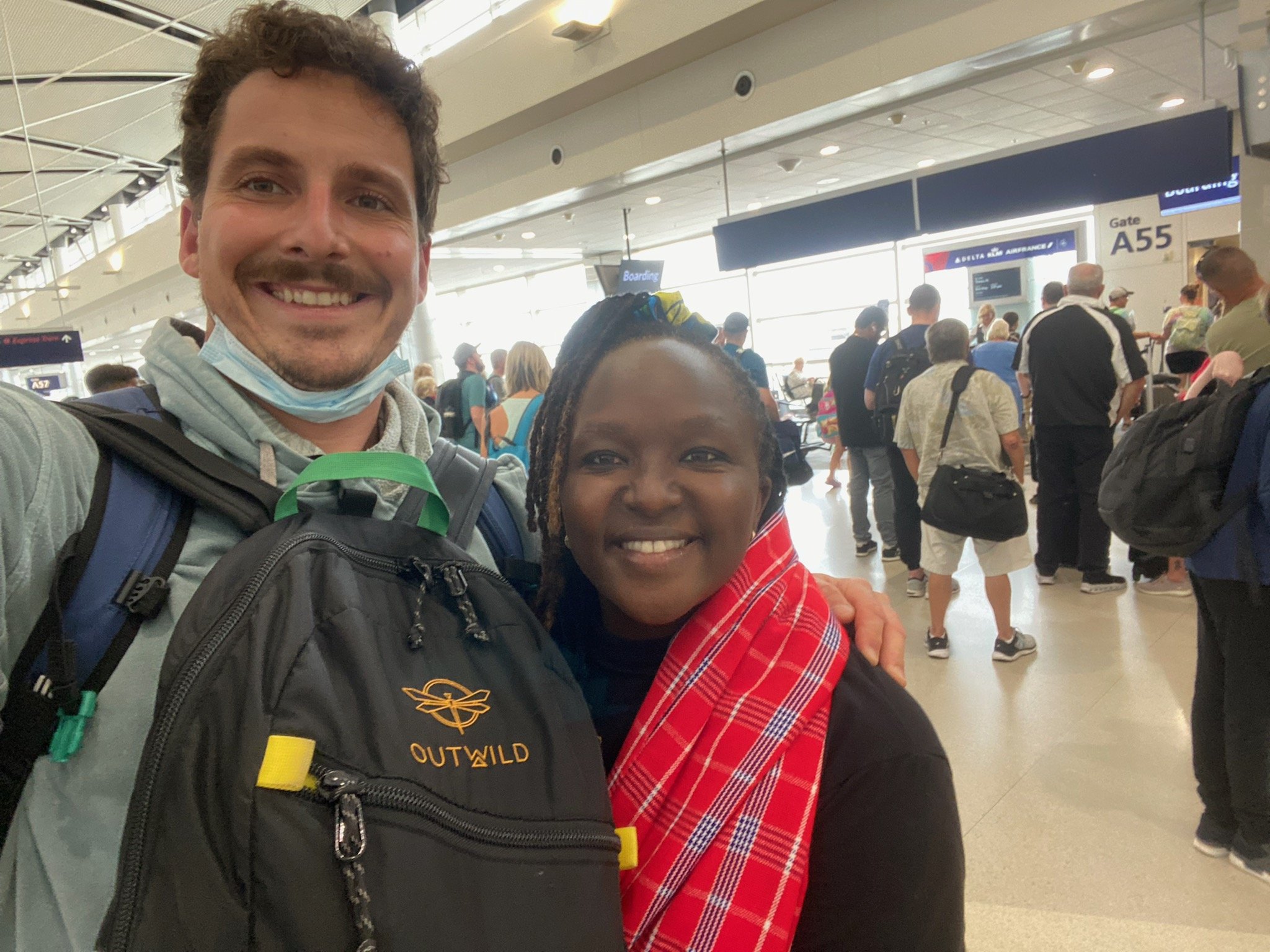 Two people smiling at an airport gate with travelers in the background.
