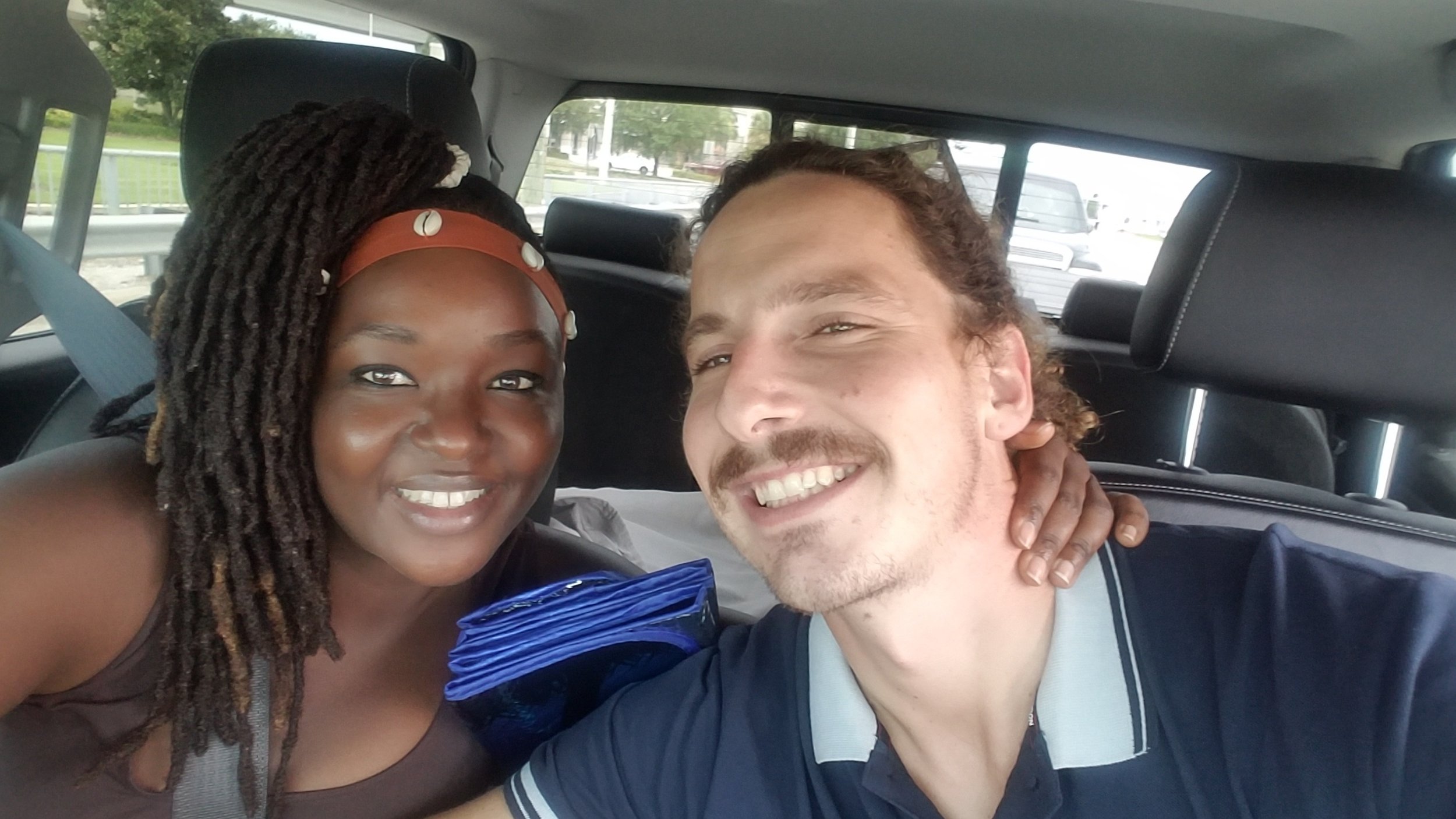 Two people smiling inside a car, one wearing a headband with shells.