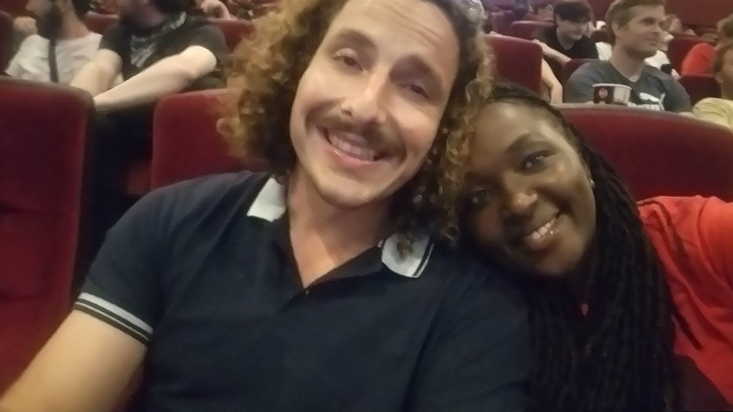 A man and woman smiling in a movie theater with red seats in the background.