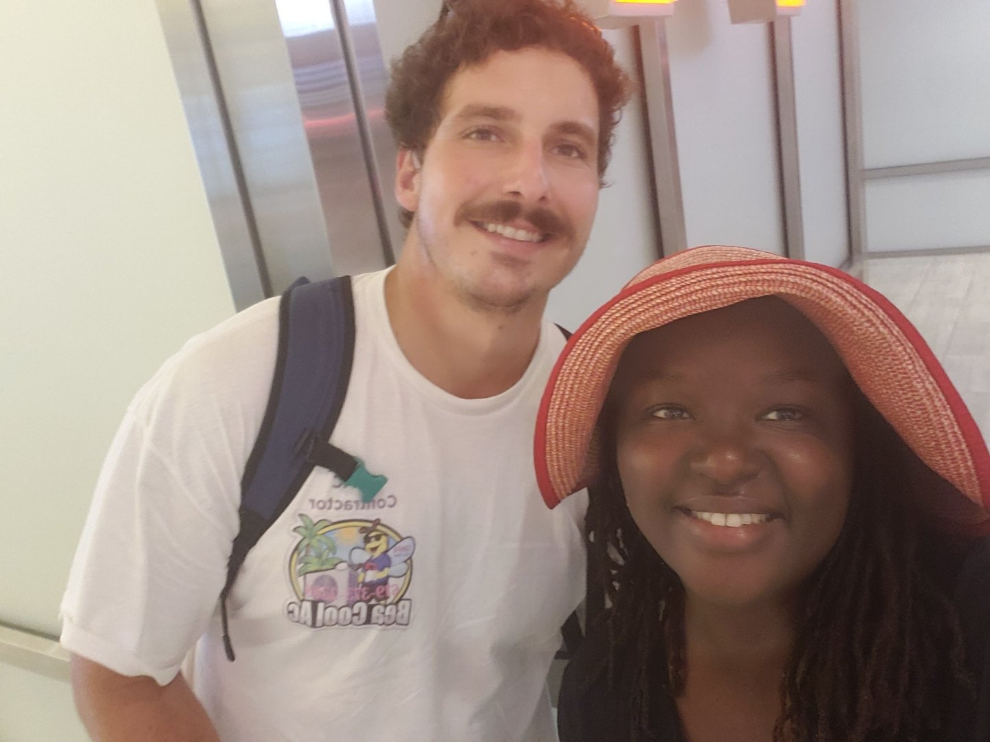 Two people smiling, one wearing a white shirt and backpack, the other a red hat, standing in a well-lit indoor setting.