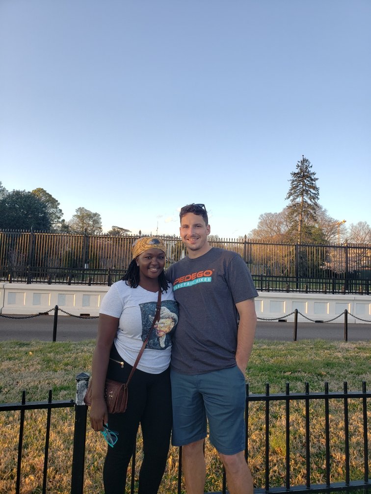 Two people standing together in front of a fence with trees and a clear blue sky in the background.