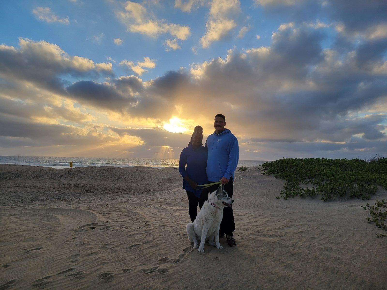 Couple with a dog standing on a sandy beach during sunset, with a dramatic cloudy sky.