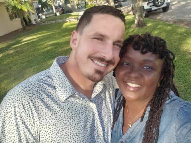 A smiling couple taking a selfie in a sunny outdoor setting with grass and trees in the background.