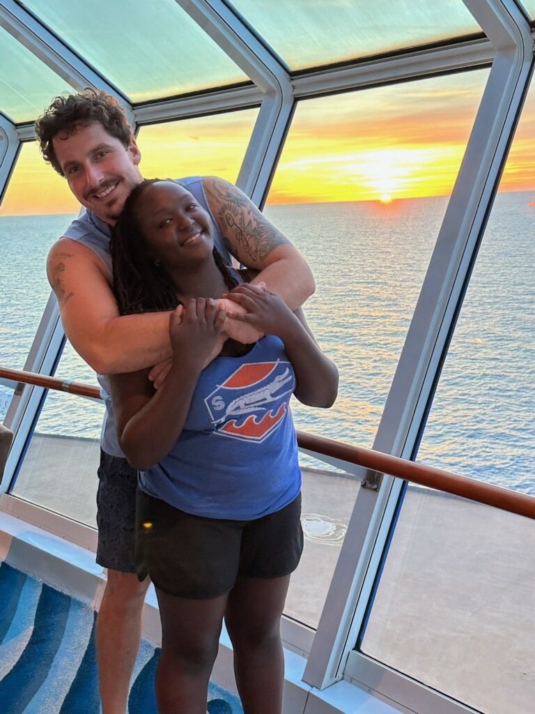 A happy couple embracing in front of a large window on a cruise ship with a sunset view over the ocean.