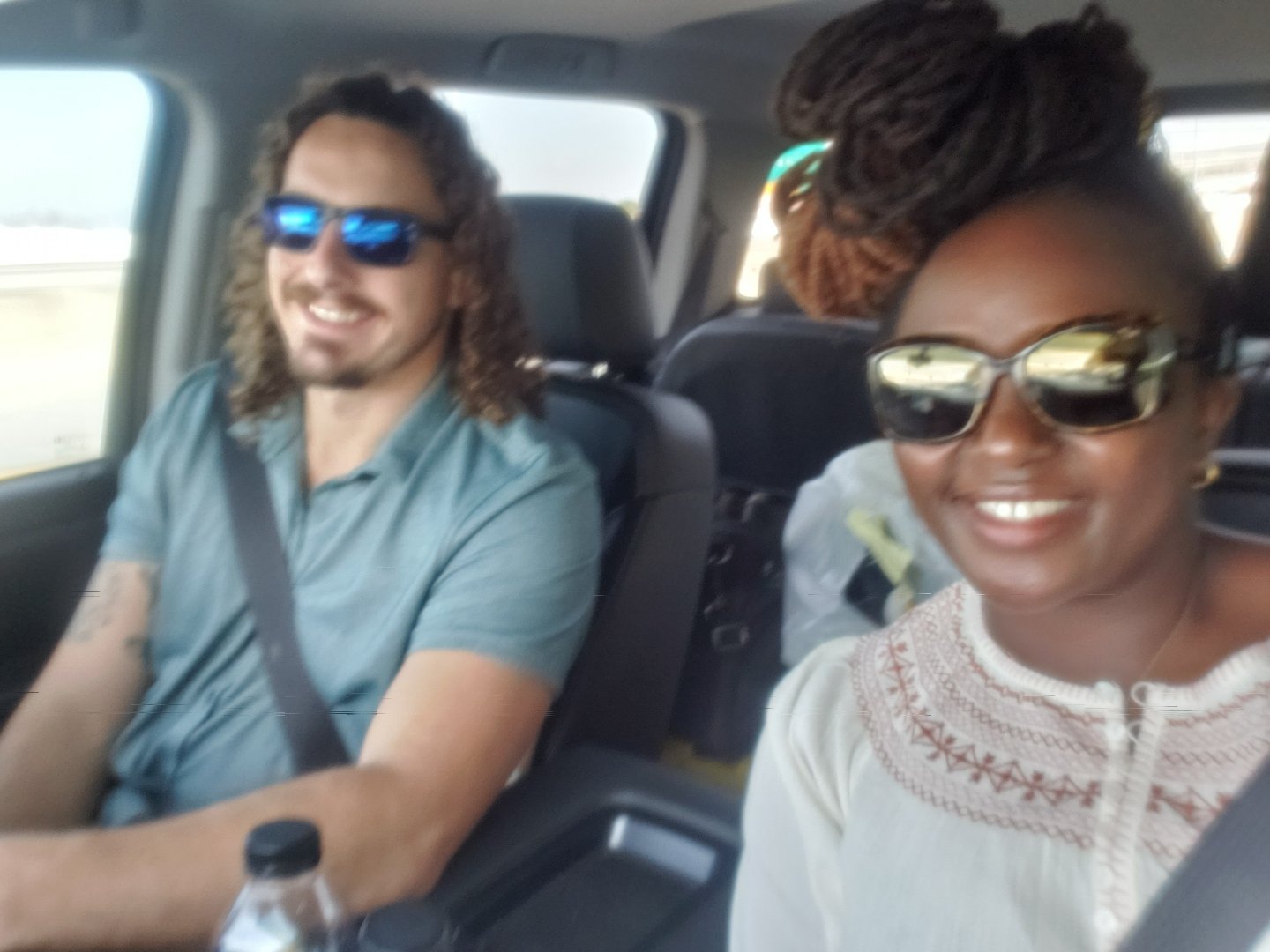 Two people wearing sunglasses smiling inside a car, with seatbelts on, during a daytime drive.