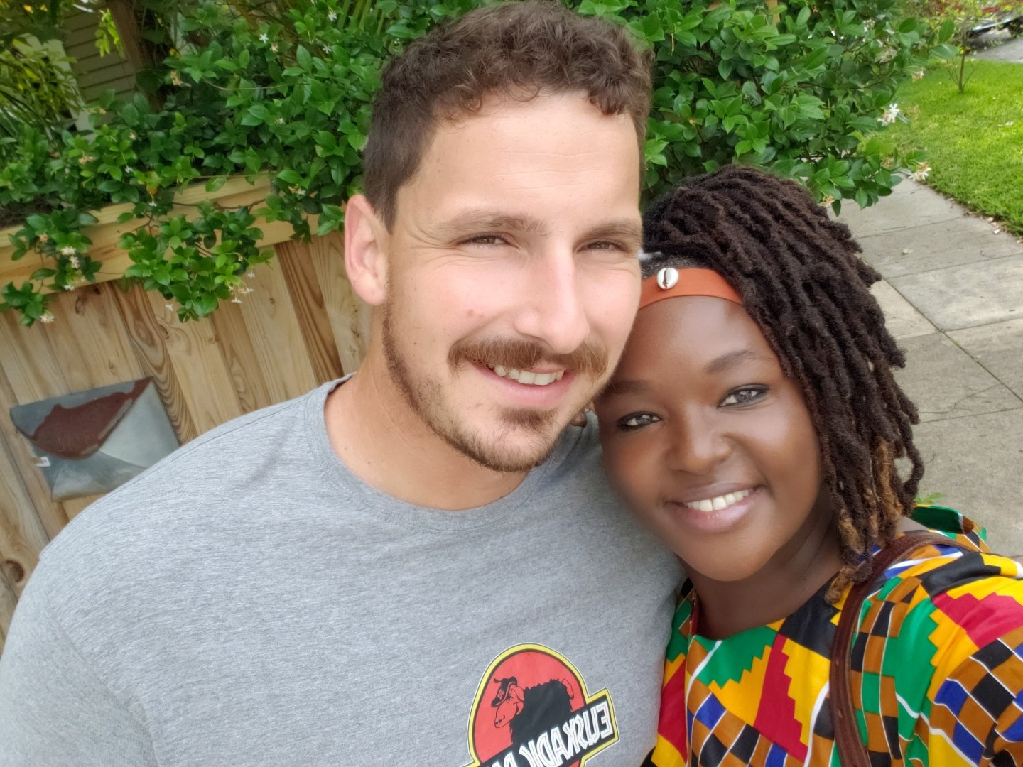A smiling couple taking a selfie outdoors in front of green foliage. The man is wearing a gray t-shirt, and the woman is wearing a colorful patterned top with a headband.