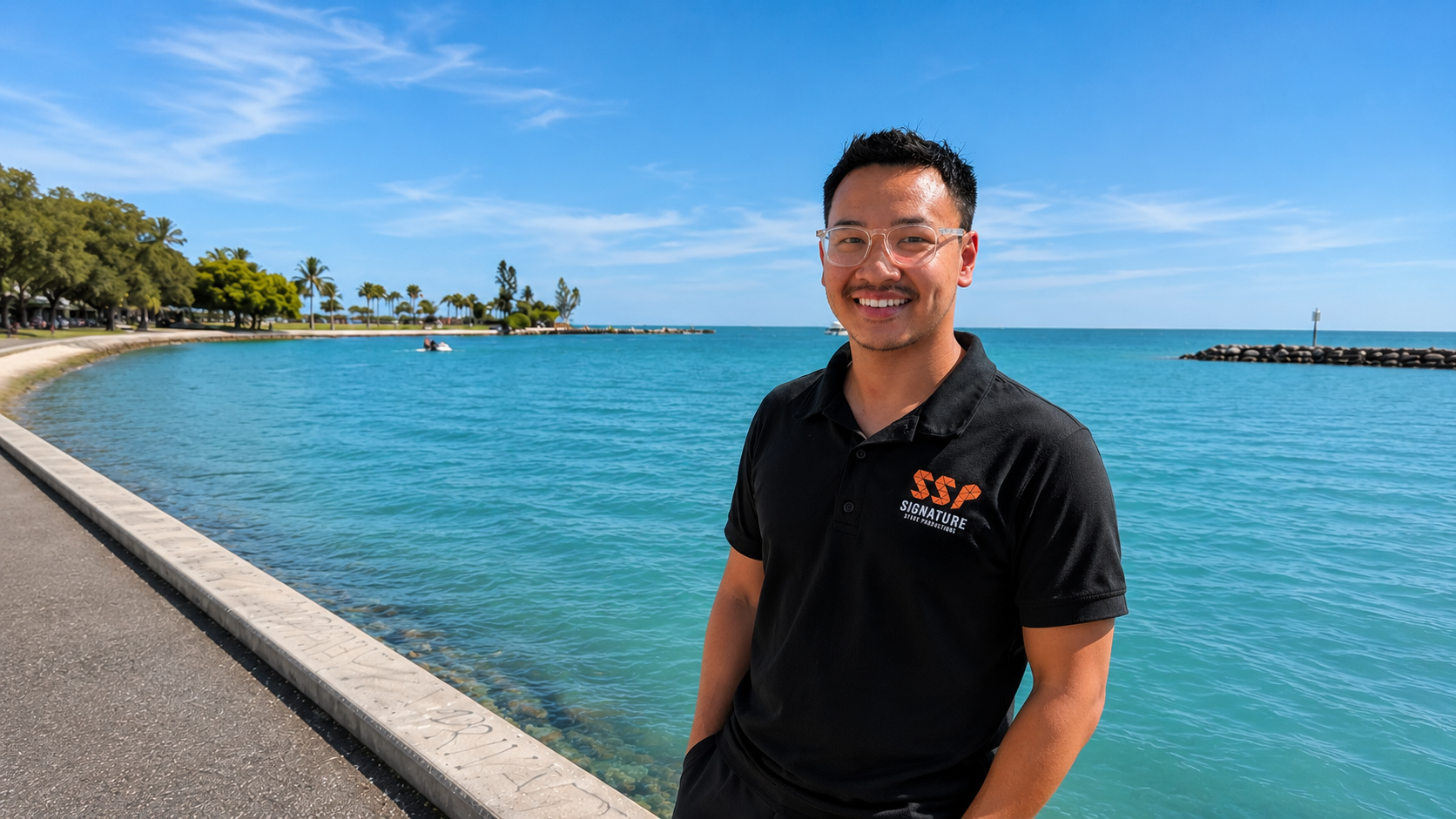 A smiling man wearing glasses and a black polo shirt with a logo stands by a waterfront with clear blue water, trees, and a partly cloudy sky in the background.