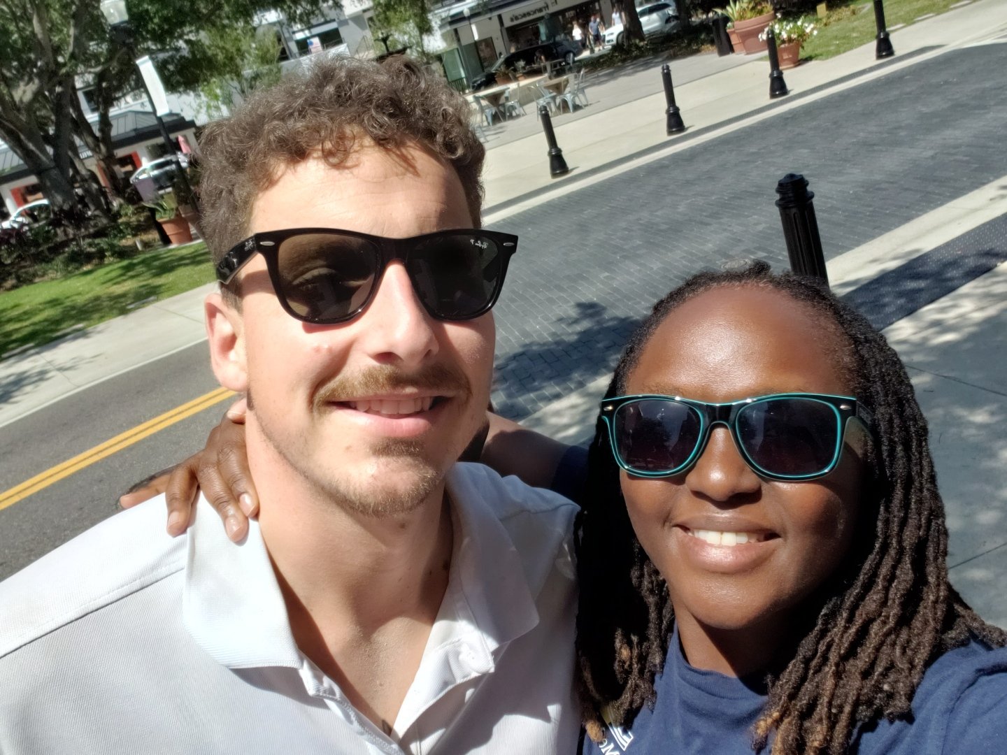 Two people wearing sunglasses smiling outdoors on a sunny day, posing for a selfie. They appear to be on a sidewalk with greenery and buildings in the background.