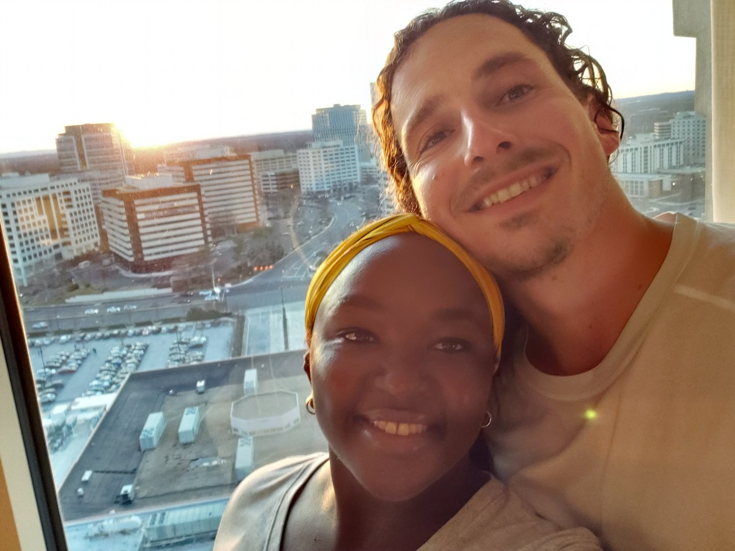 Two people smiling in front of a cityscape, seen through a window with buildings and sunset in the background.