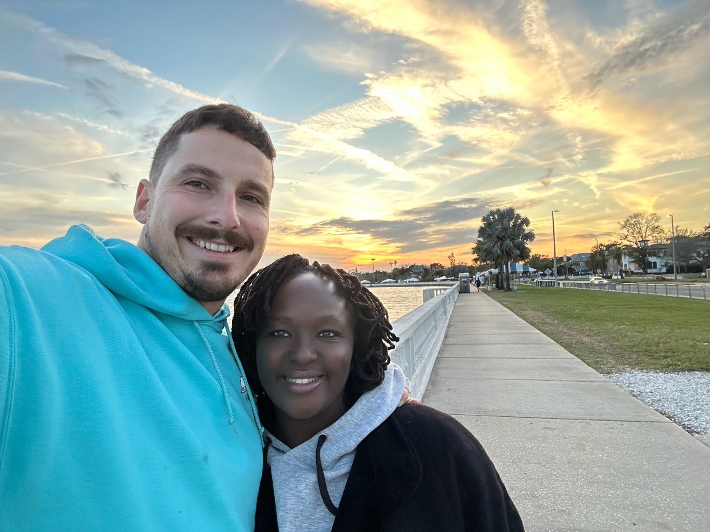 A couple smiling for a selfie near a waterfront at sunset, with a walkway and palm trees in the background.