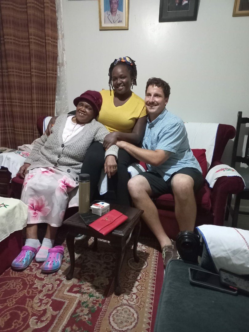 Three people sitting closely together on a couch in a living room. An older woman wearing a maroon hat, patterned skirt, and sweater sits on the left. A younger woman in a yellow shirt is in the middle, and a man in a blue shirt and shorts is on the