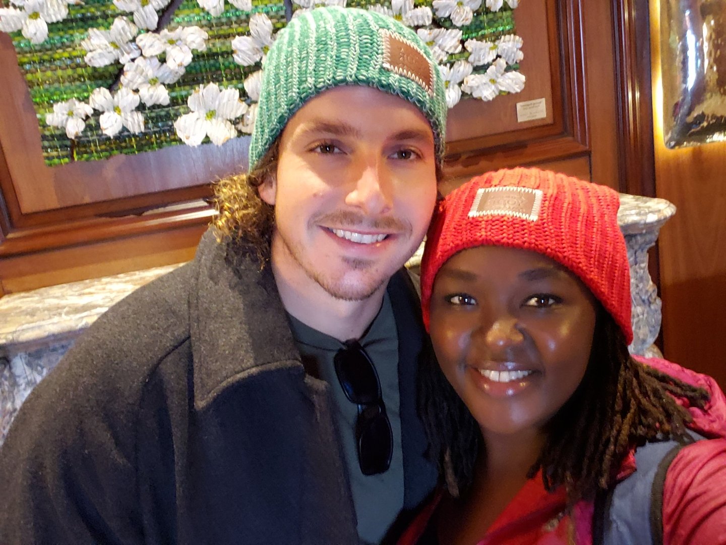 A smiling couple wearing knitted beanies, standing in front of a decorative floral wall art.