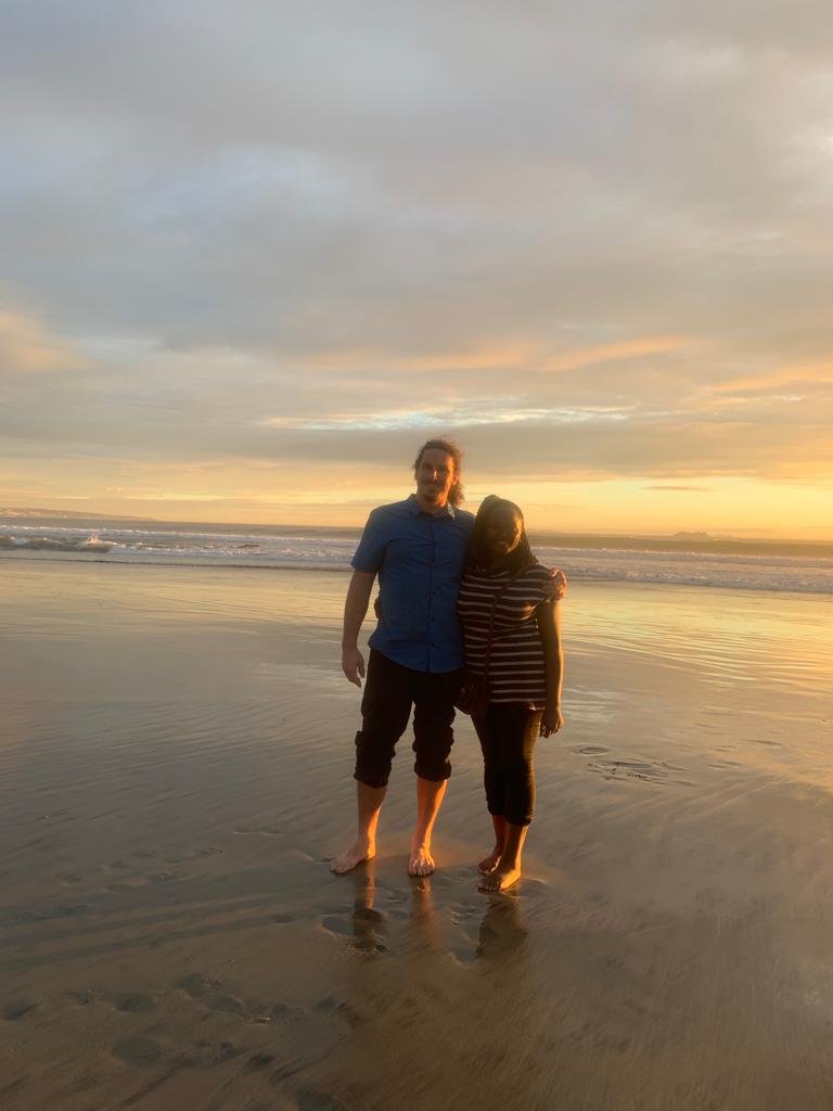 A couple standing barefoot on the beach during sunset, with the ocean in the background.