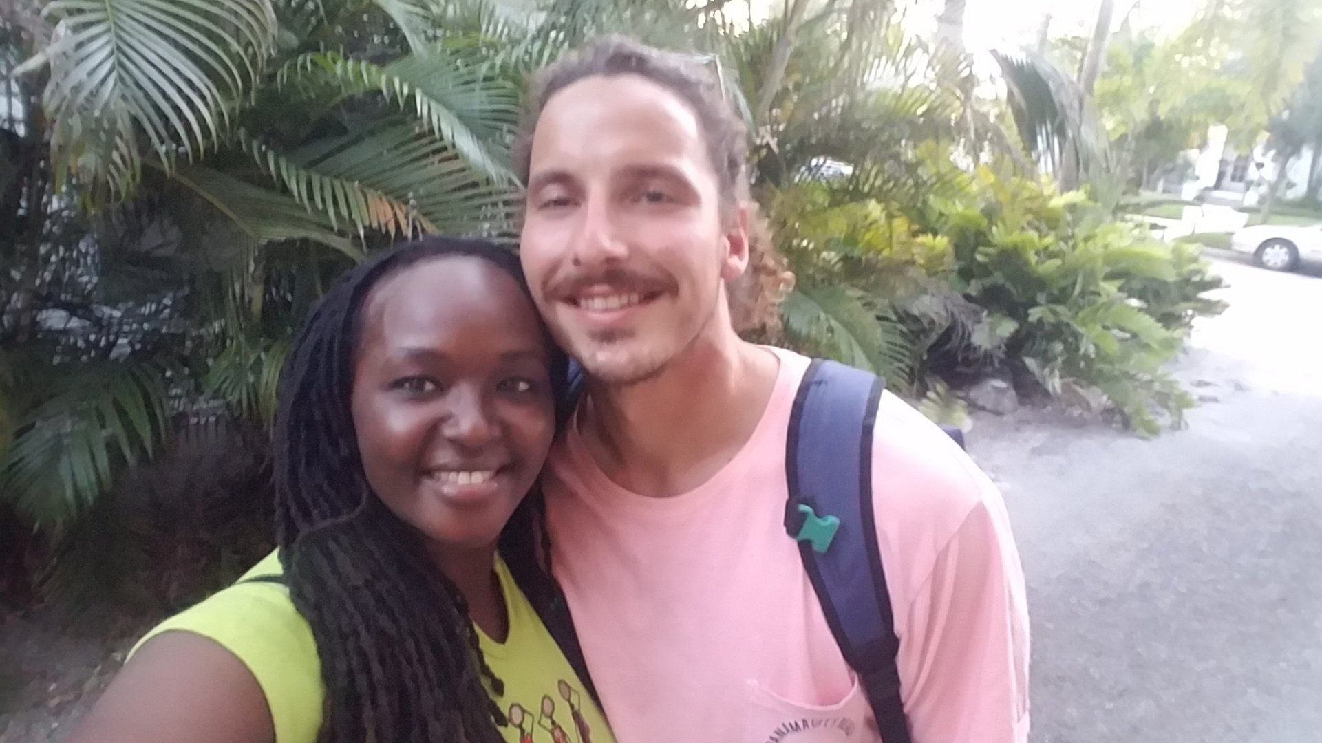 Two people smiling outdoors with tropical plants in the background.