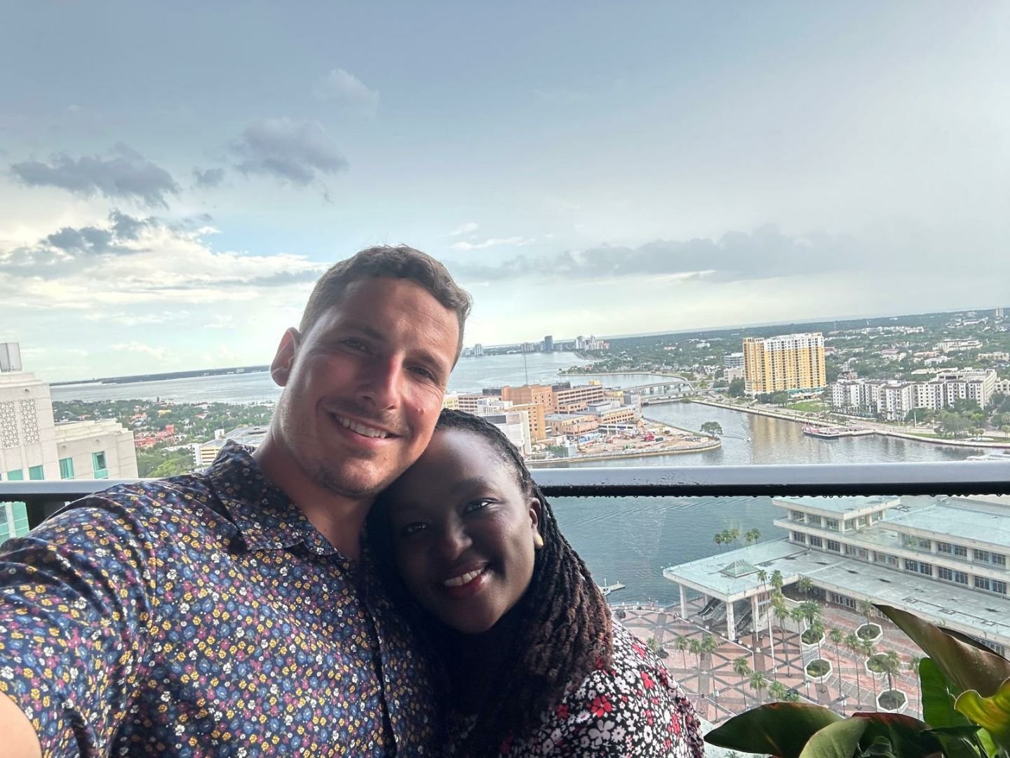 A couple taking a selfie on a balcony with a city and river view in the background.