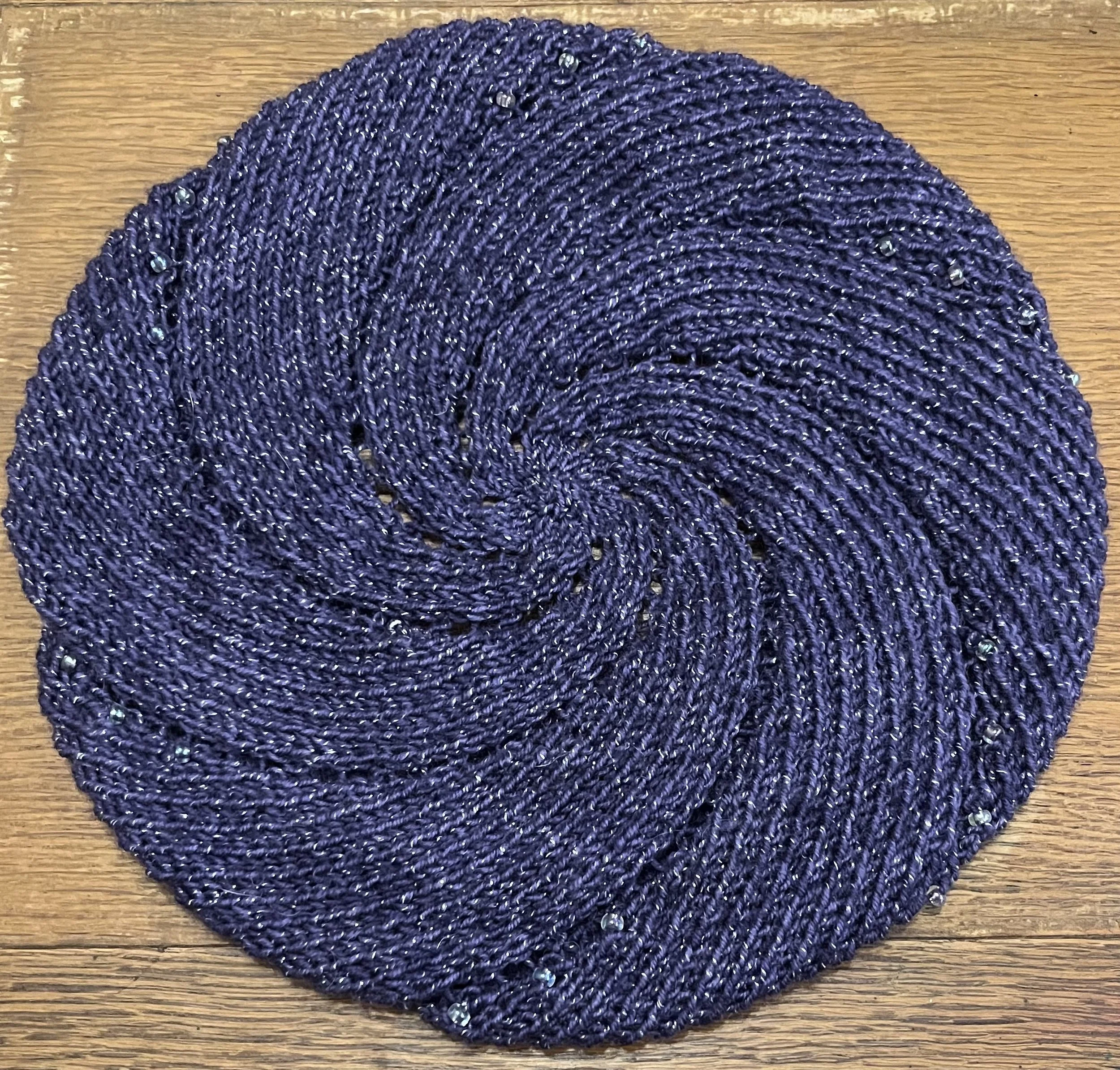 A knit beret made of purple glittery yarn. The beret is made of 6 spiraling sections. There are glass beads along the edges of each section.