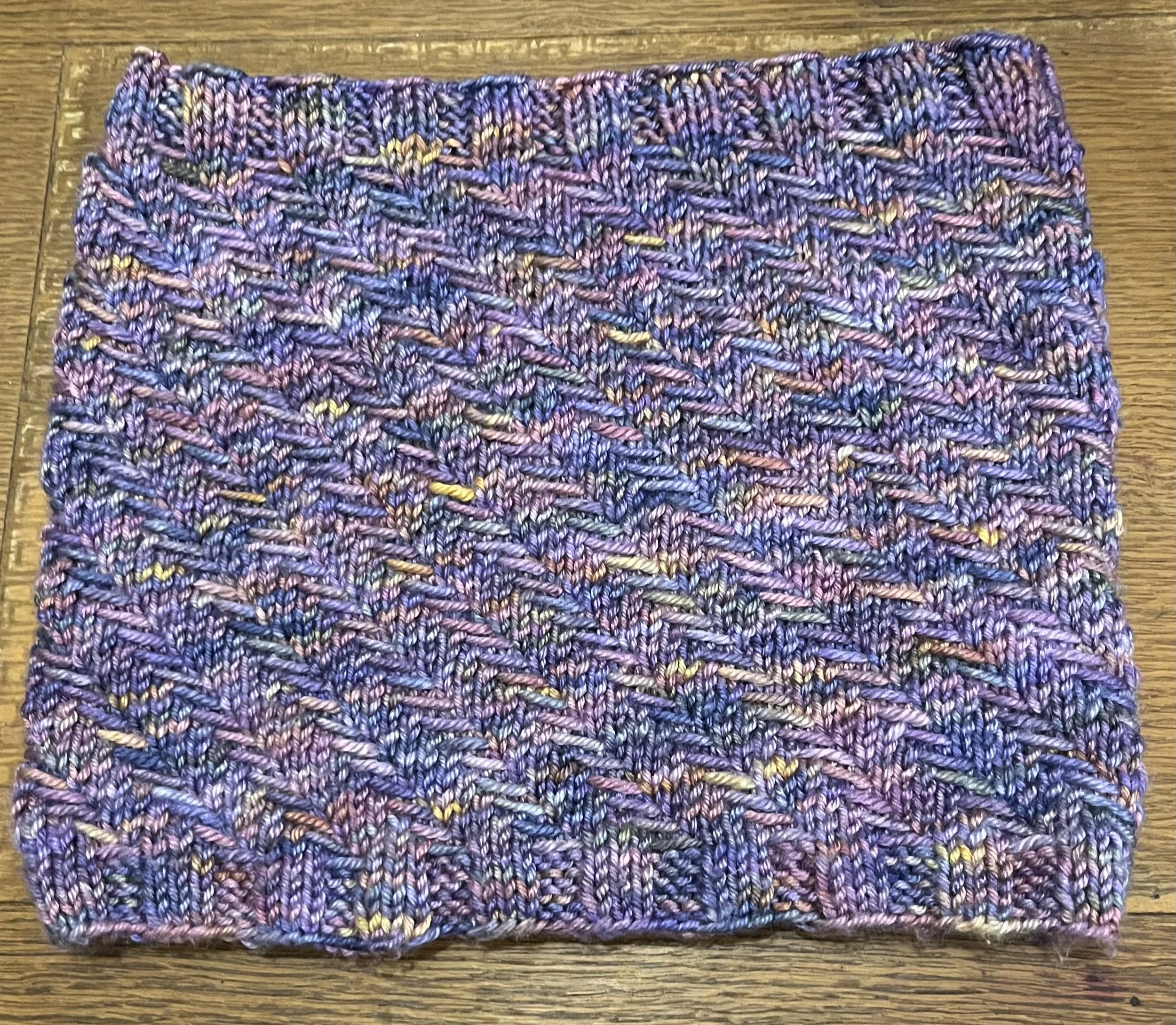 A knit cowl in a purple, blue and orange color way. there are dashes of horizontal slipped yarn in a spiraling pattern.