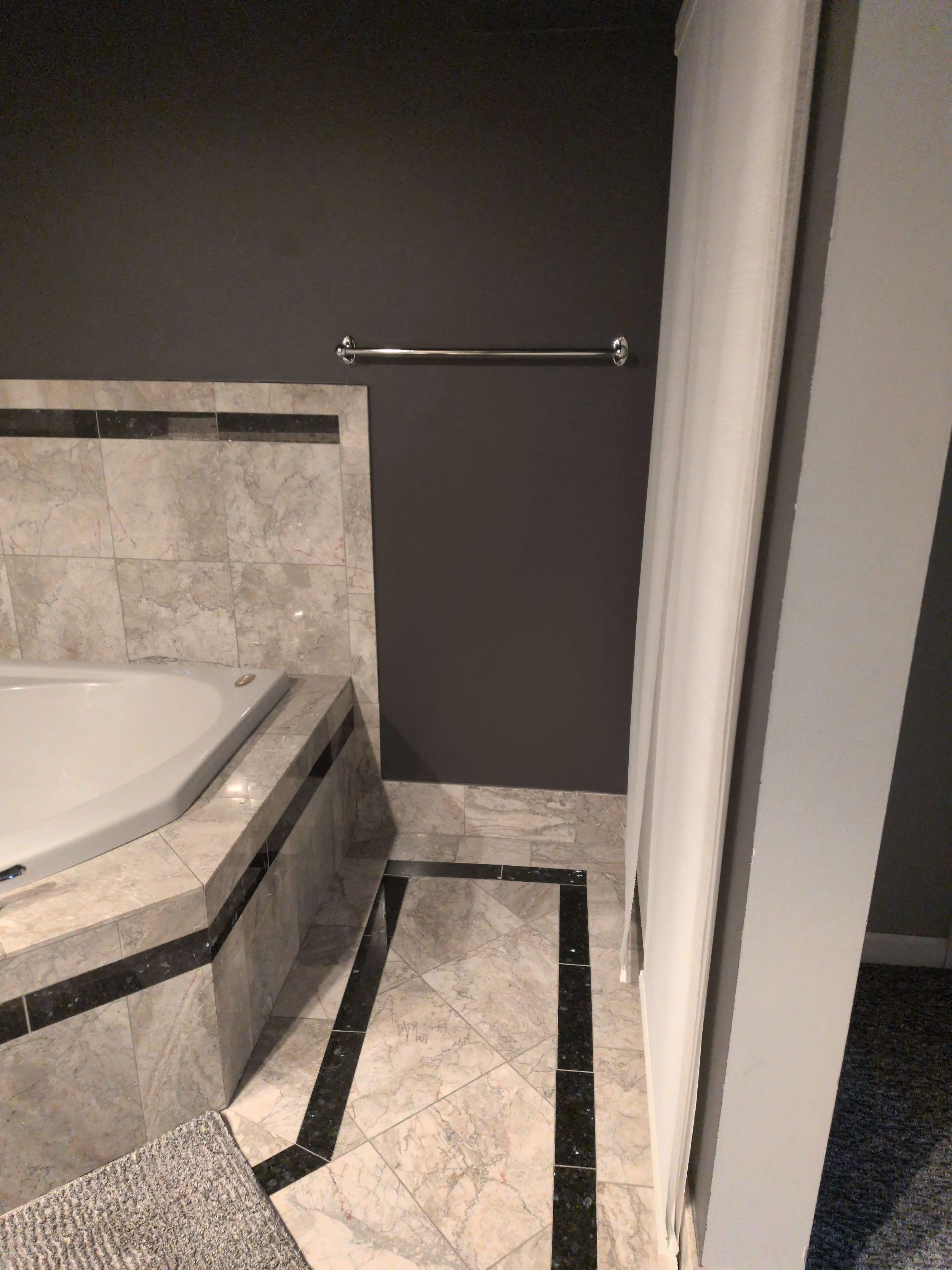 Primary focus of this image is of a narrow strip of gray wall. It is so narrow, the bar to hold a full size towel extends over the tub.