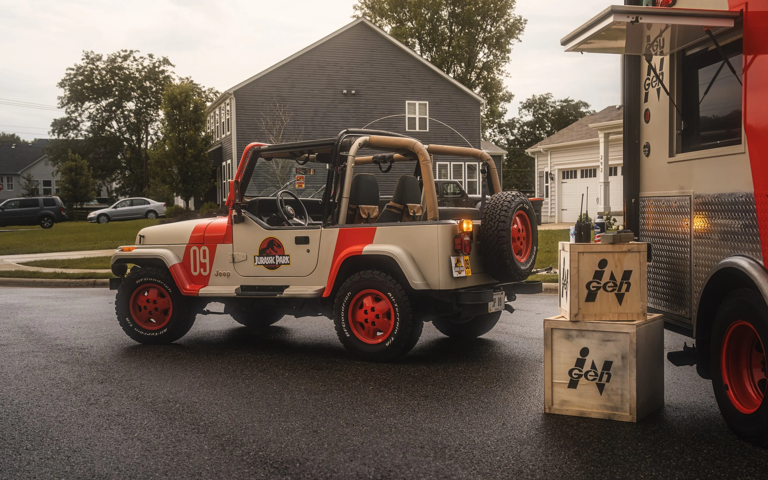 A Jurassic Park-themed Jeep with red wheels and a number 09 decal is parked next to a food truck or trailer on a wet street in a residential neighborhood.