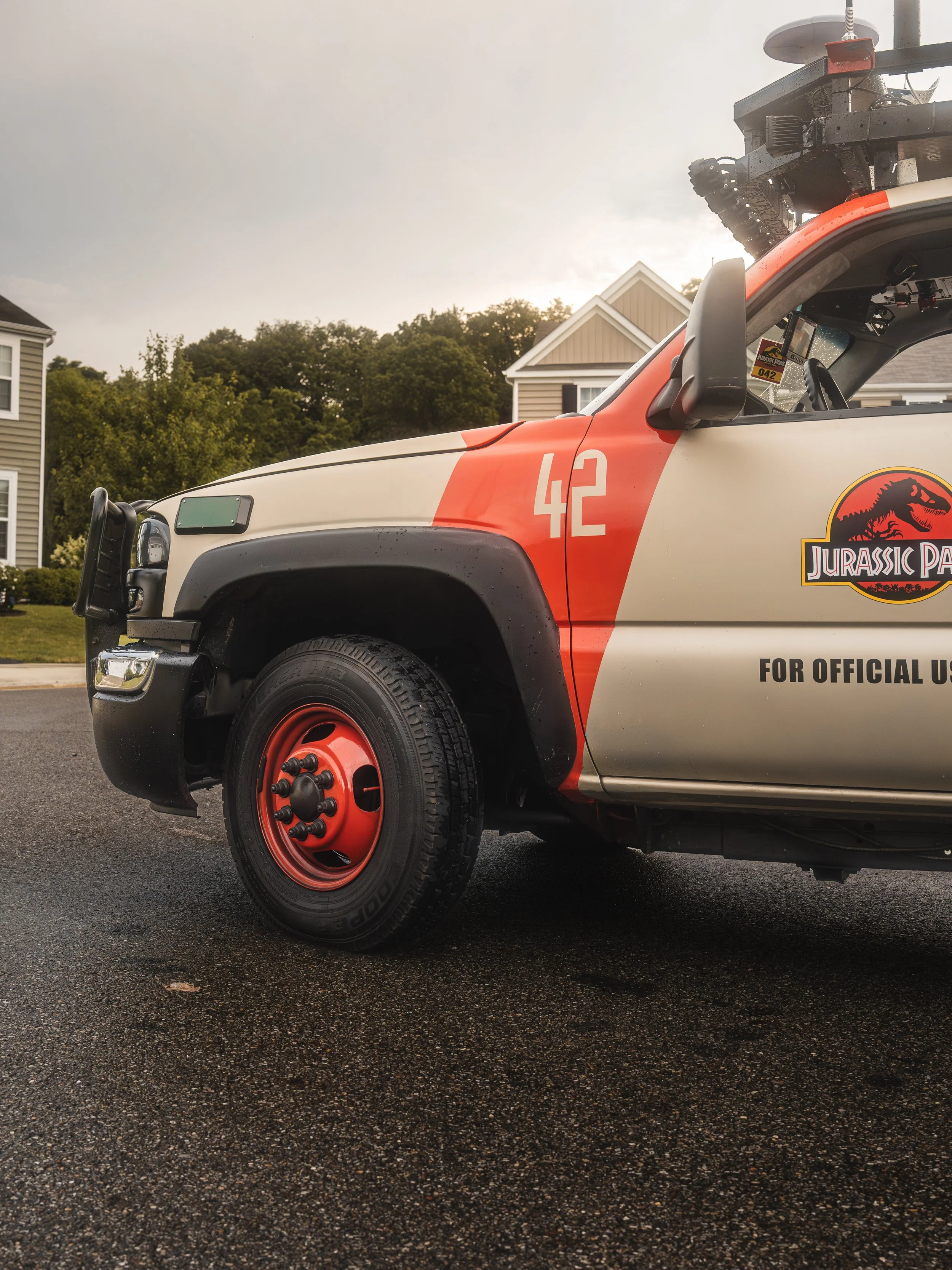 Close-up of a police vehicle with a Jurassic Park logo decal on the side, featuring a white background with red accents and the number 42. The vehicle is parked on a wet asphalt road in a residential area.