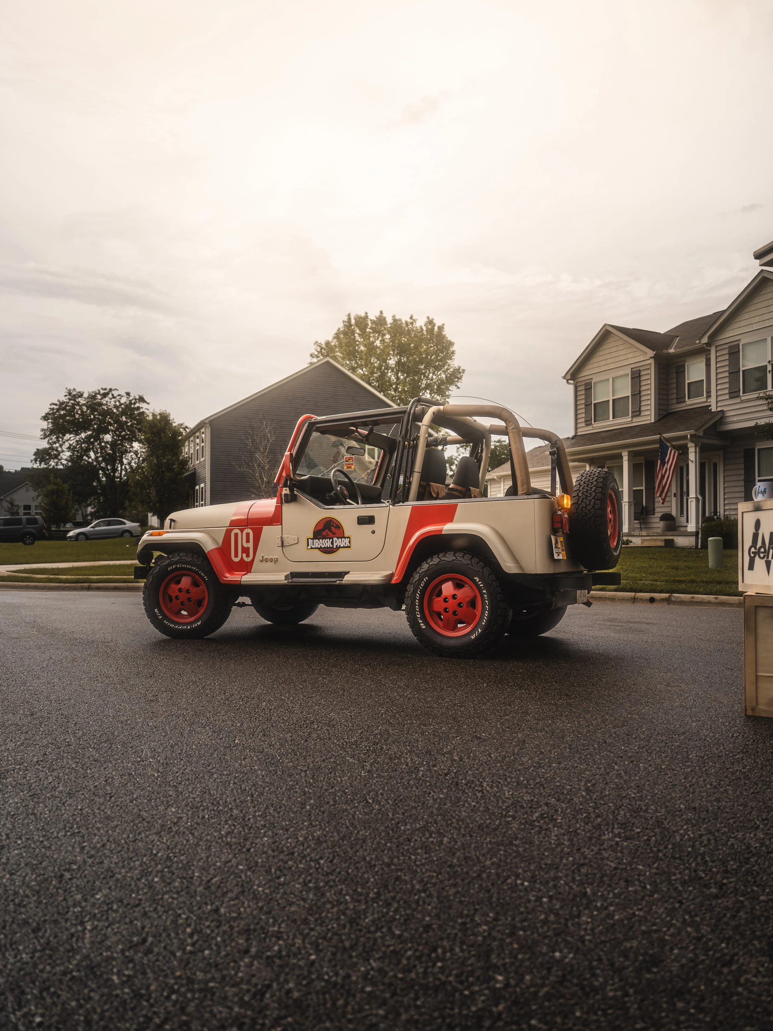 A Jurassic Park-themed toy Jeep with red wheels and a beige body parked on a residential street during a cloudy day.