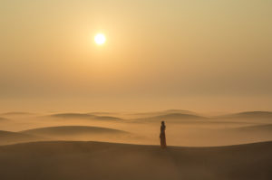 A lone person stands in a the desert sand dunes at sunset.