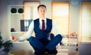Man meditating in lotus pose, wearing a blue suit on his office desk.