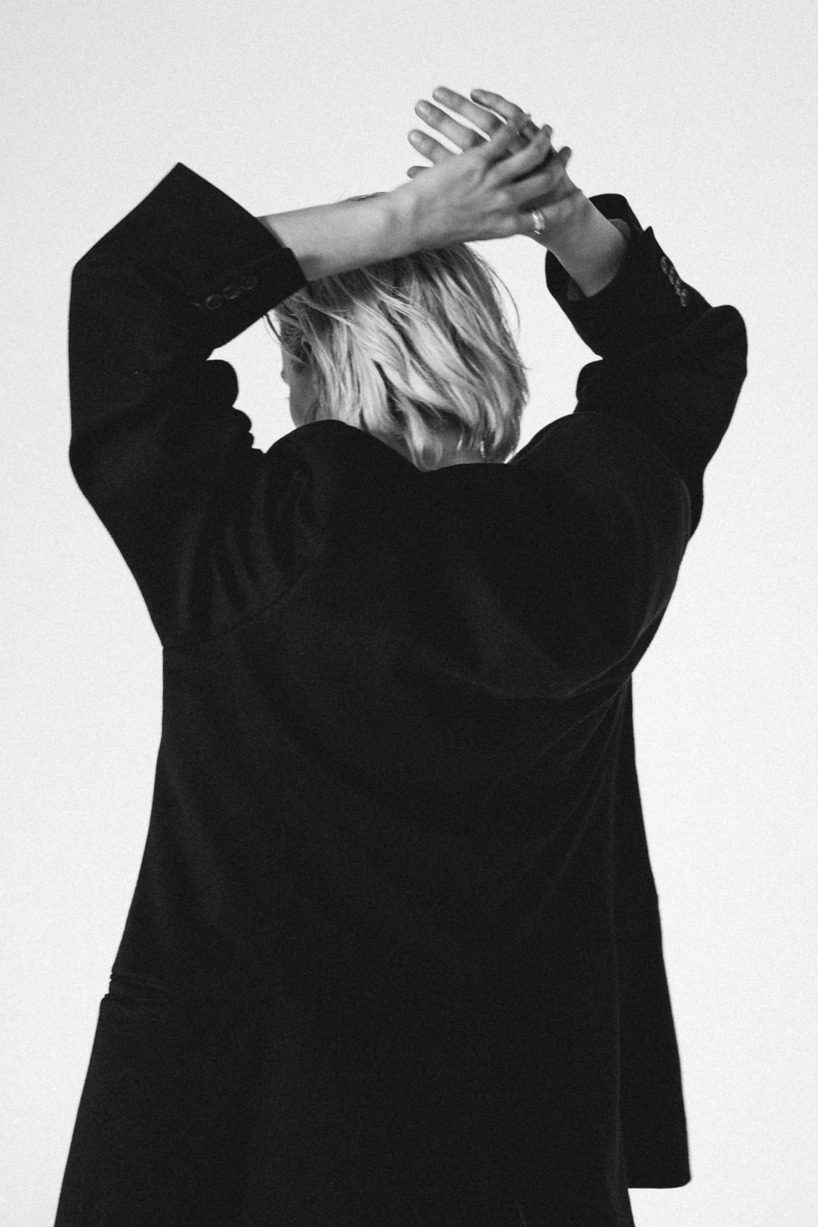 Back view of a person with short, wavy blonde hair, wearing a black blazer, with arms raised above their head, hands clasped together.