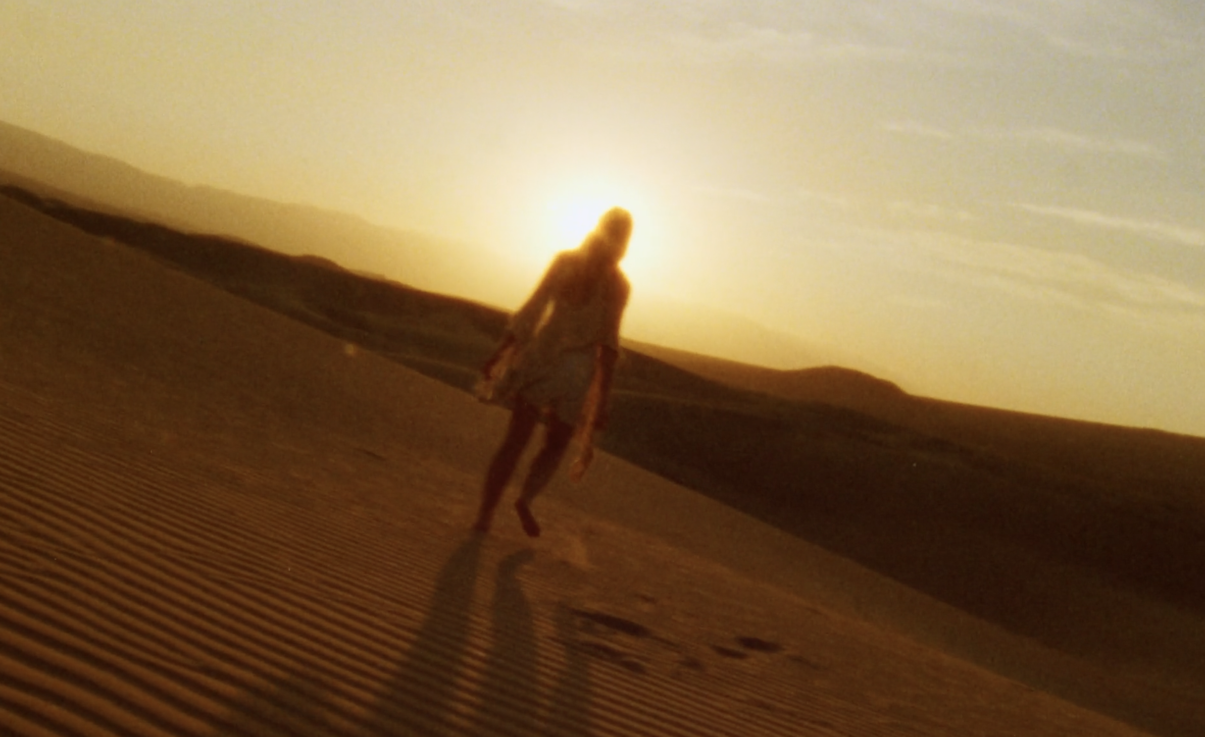 A person walking in a desert at sunset with dunes in the background.