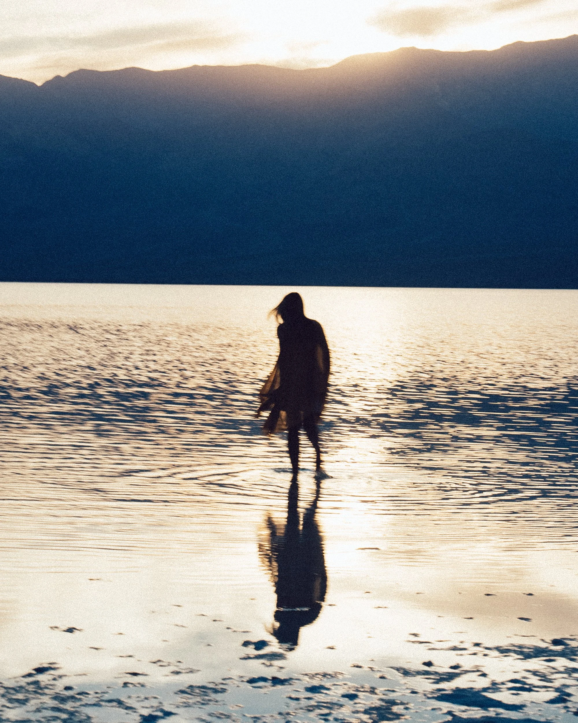 A silhouette of a person standing in shallow water at sunset, with mountains in the background and their reflection visible in the water.