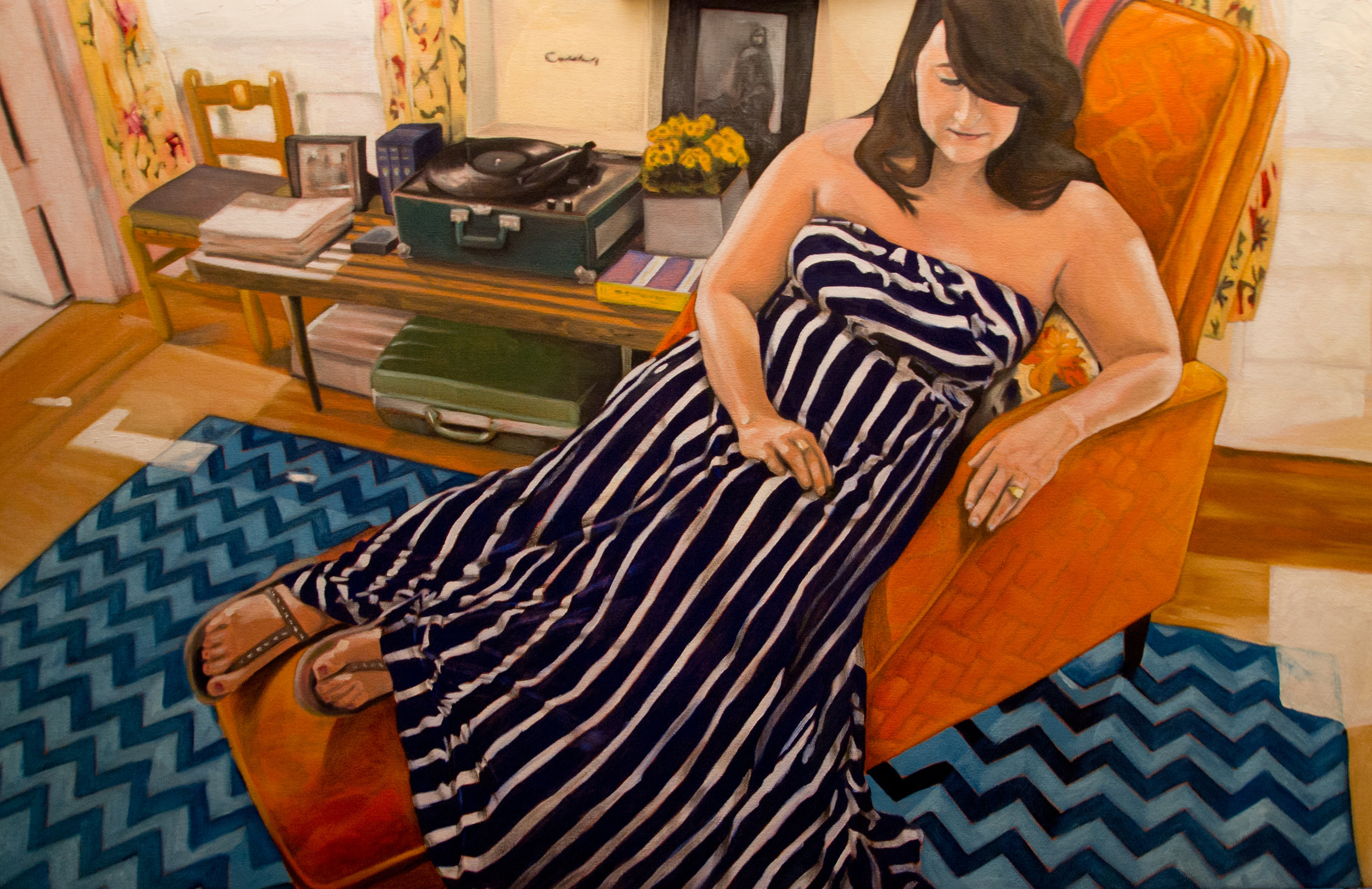 A woman in a striped strapless dress lounging on an orange armchair in a room with a wooden floor, blue and white chevron rug, and a wooden side table with books, a turntable, and decorative items.