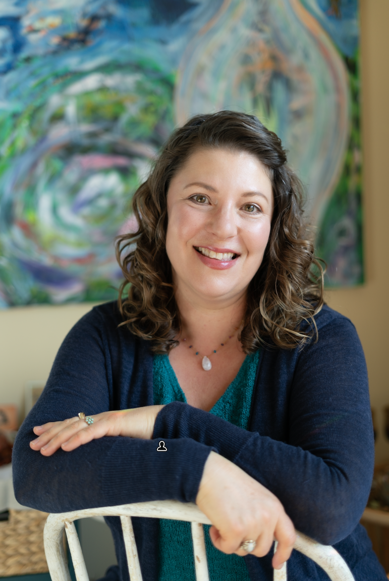 A woman with curly brown hair in a navy cardigan sits in front of an abstract painting