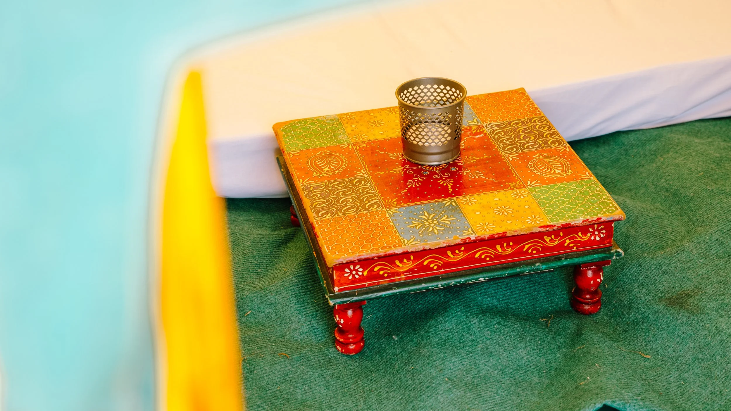 Decorative multicolored table with a metal candle holder on top, placed on green carpet with a white wall background.