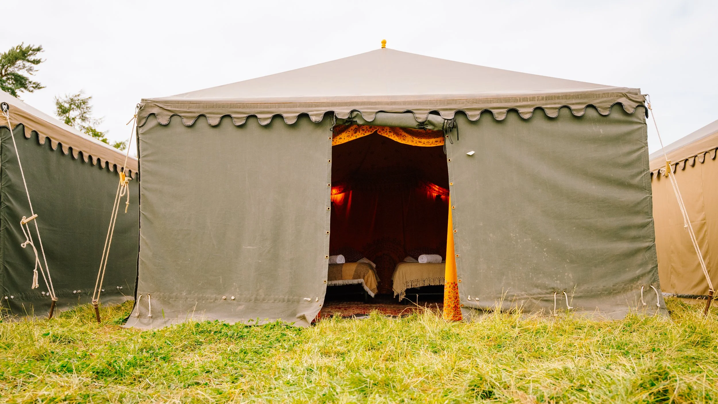 A large beige and green camping tent with an open door revealing two beds inside, set on grass outdoors under a cloudy sky.