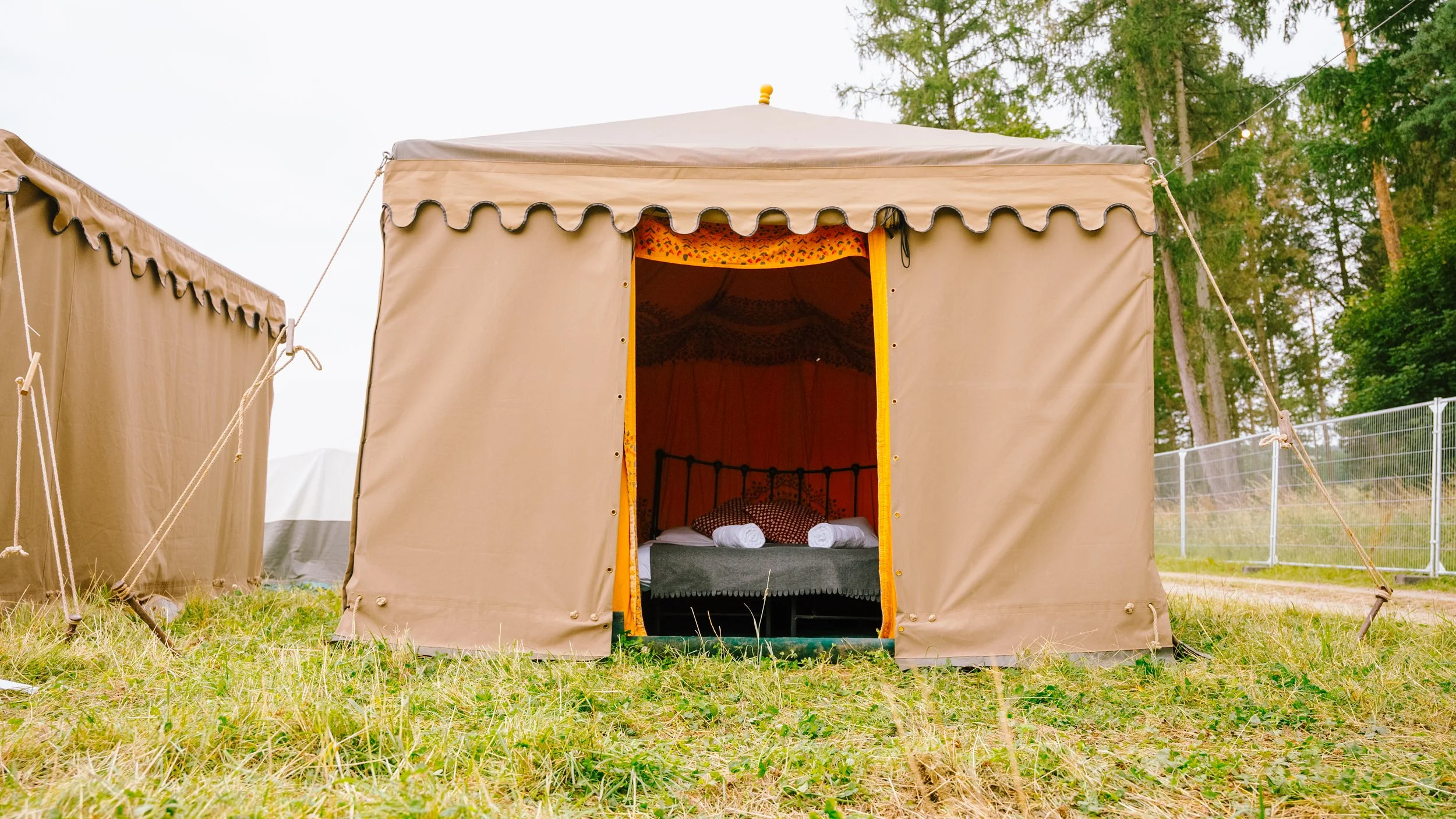 View of a beige canvas tent with an open door revealing a bed with pillows inside, set on grassy ground with trees in the background.