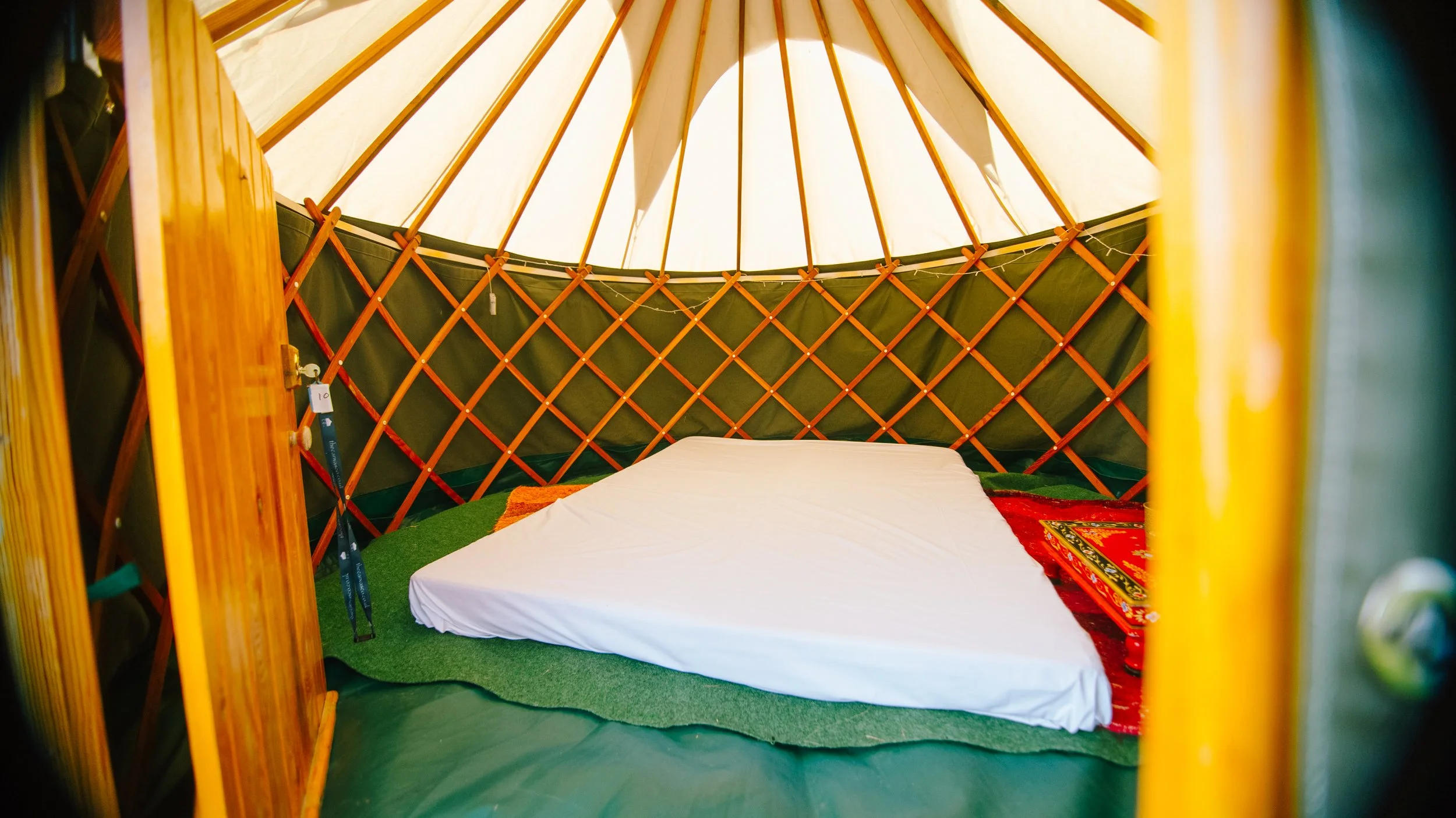 Inside a yurt with a bed on a green rug and colorful decorative pillows, wooden lattice walls, and a canvas roof.