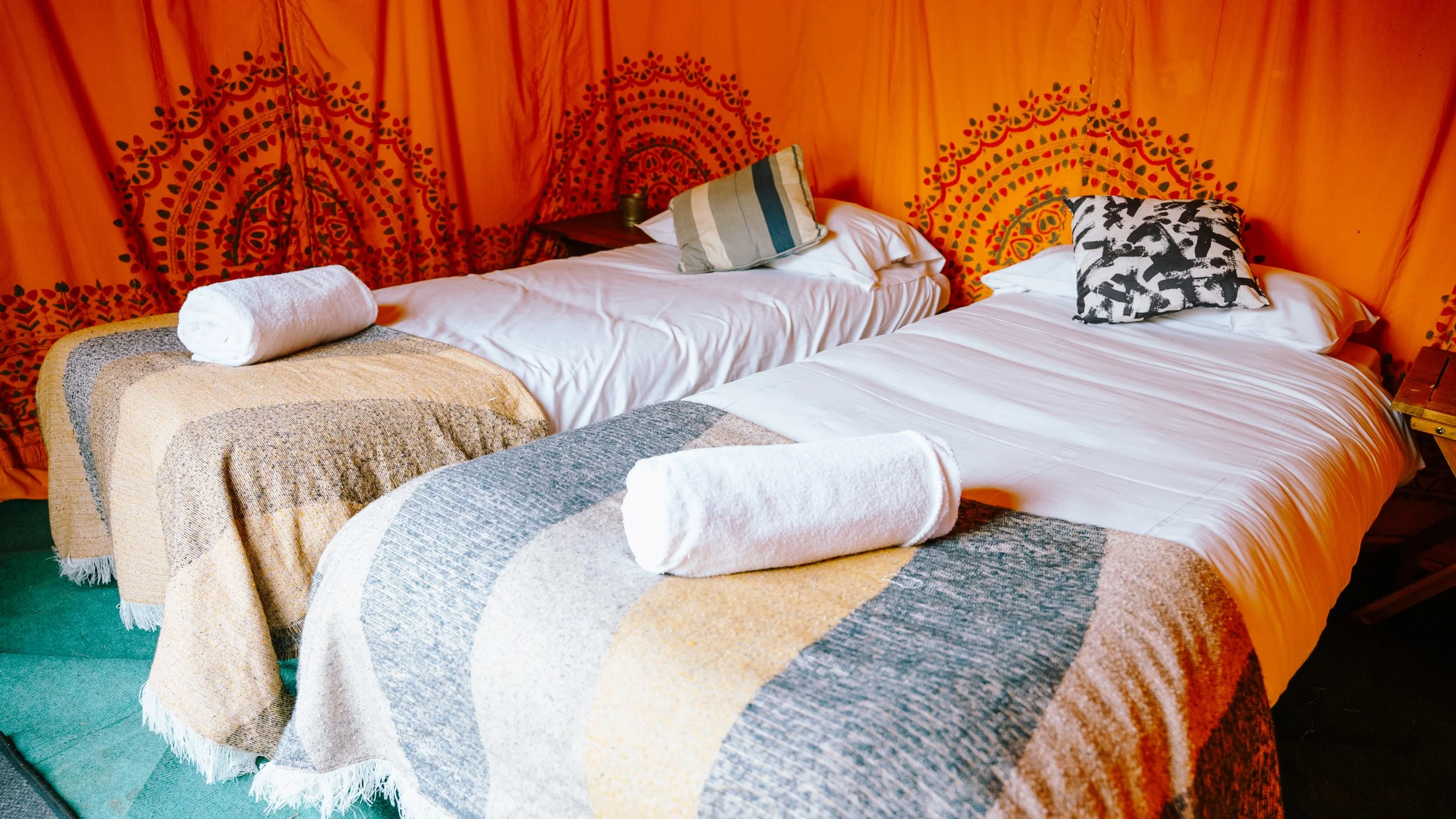 Two beds with white sheets, colorful woven blankets, rolled white towels, and patterned pillows, against an orange wall with decorative designs.