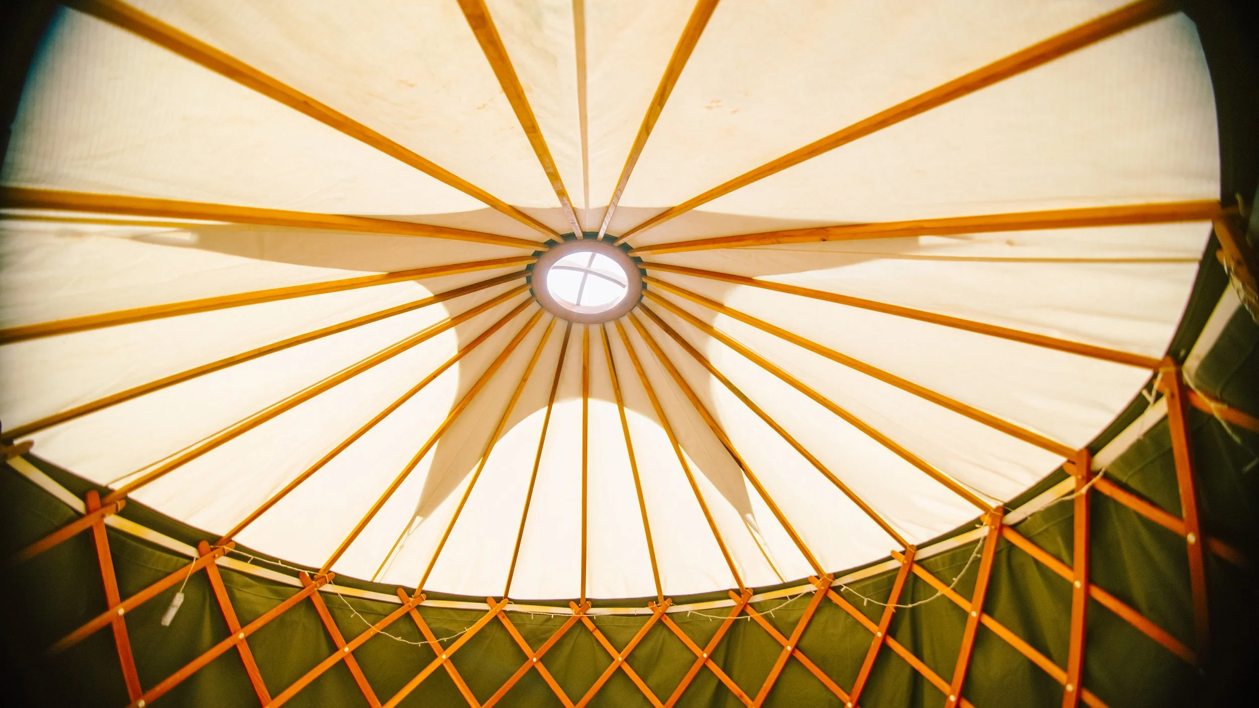 Upward view of a beige tent canopy with orange support beams, with a circular opening at the top allowing light to shine through.