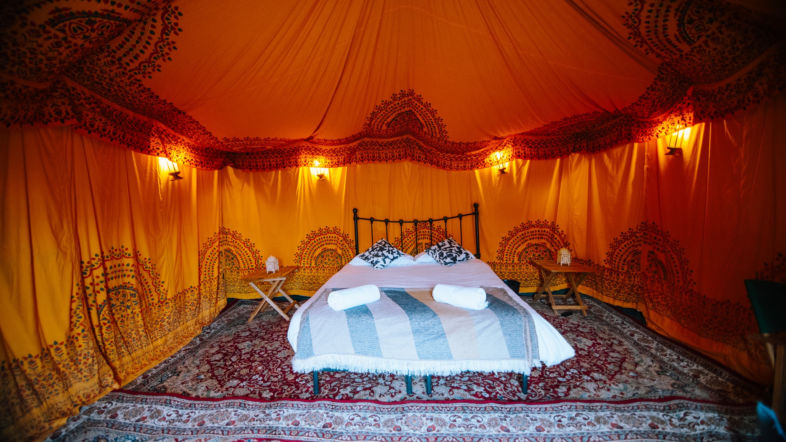 Interior of a luxurious bed inside a Moroccan-style tent with orange fabric walls and ceiling, patterned rugs, and black metal bed frame, decorated with pillows and towels.
