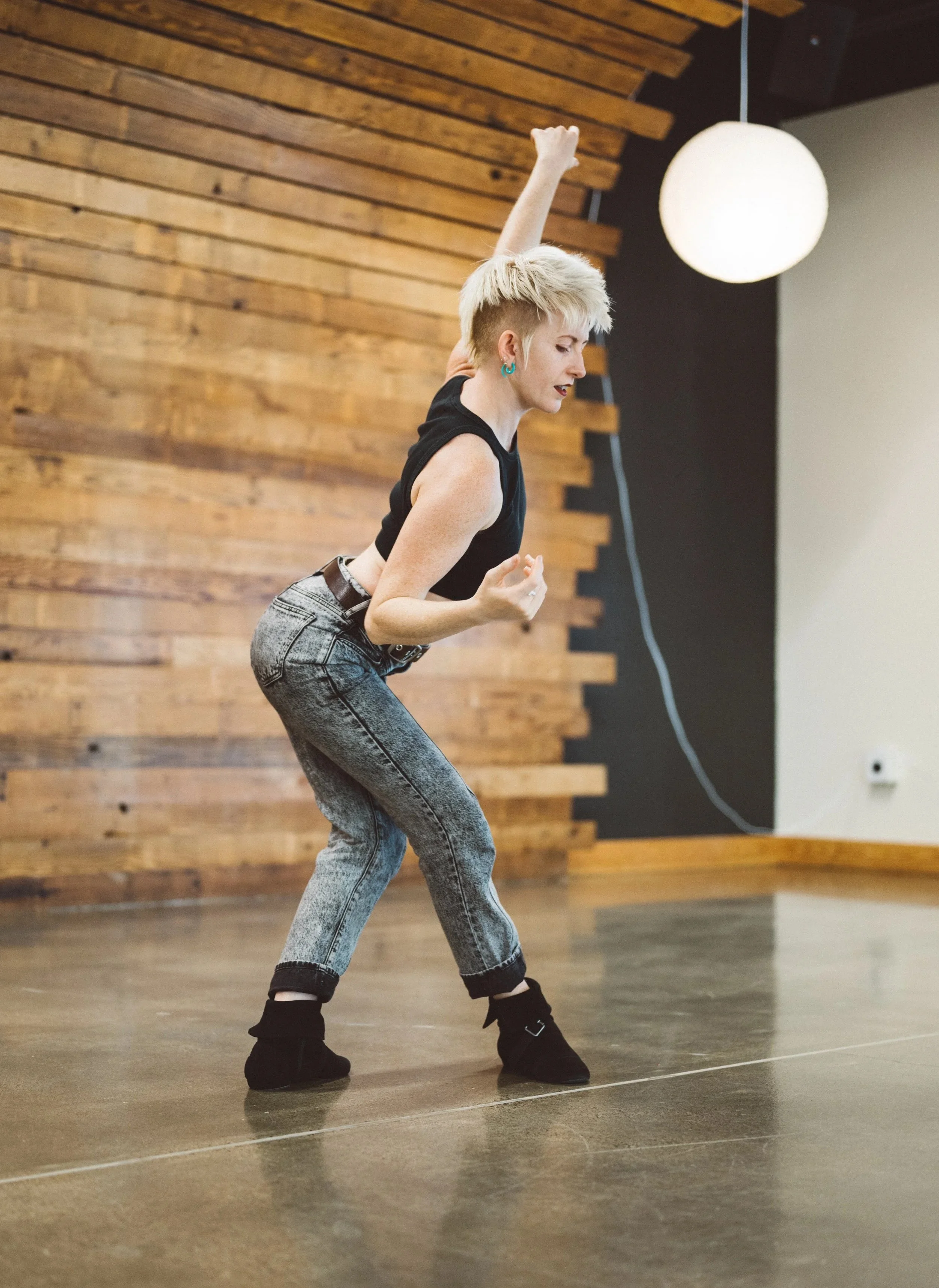 Ally Yancey dancing in a studio, demoing for a queer somatic movement class. She is wearing grey jeans and a black crop top, and her arm reaches up expressively.